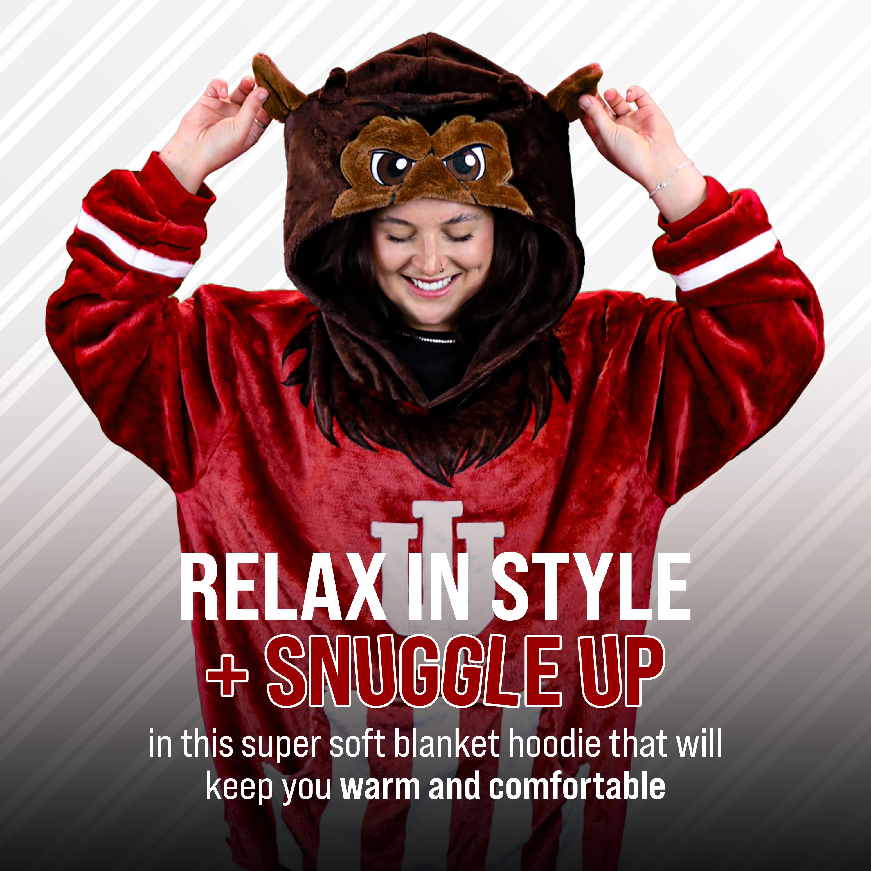 A person wearing a plush red and brown Bison mascot hooded blanket costume poses with hands on the hood ears against a gray diagonal-striped background. Large text reads 'RELAX IN STYLE + SNUGGLE UP' and smaller text below describes comfort.